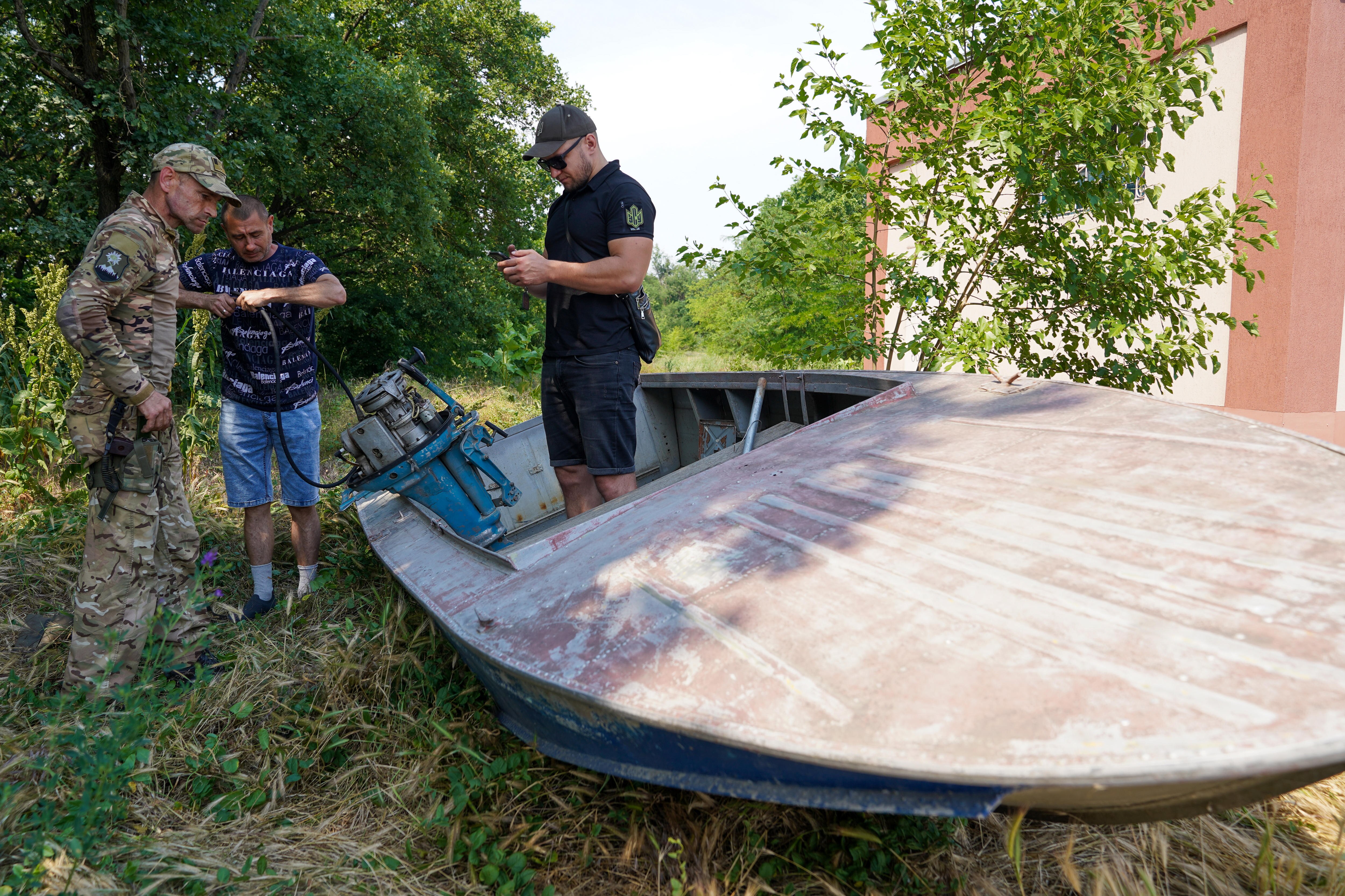 Three men stand around a boat. One looks at a phone while the other two tinker with the motor