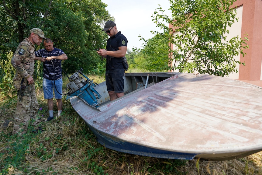 Three men stand around a boat. One looks at a phone while the other two tinker with the motor