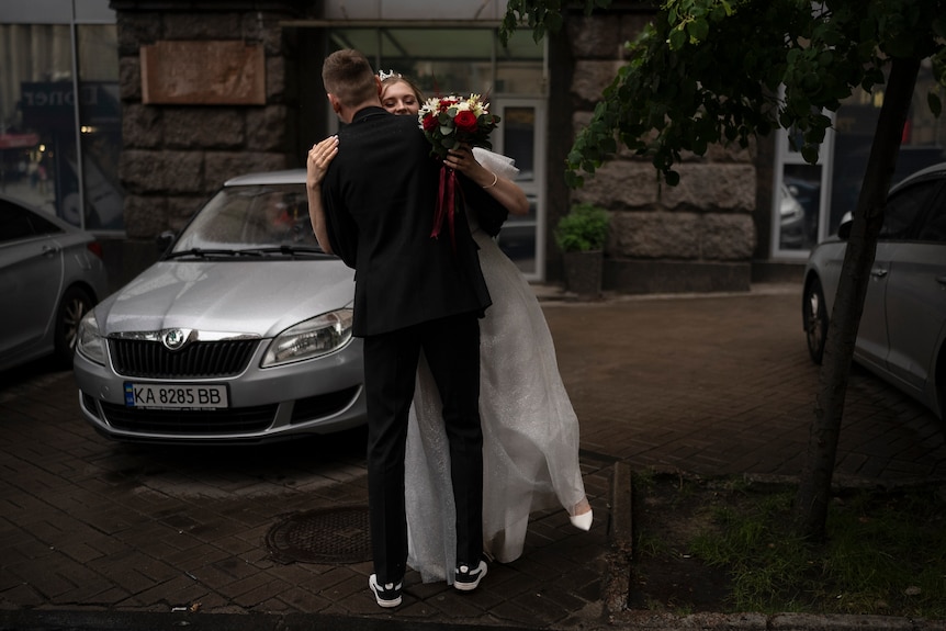 A young man in a suit and a young woman in a wedding dress embrace as they dance on the street