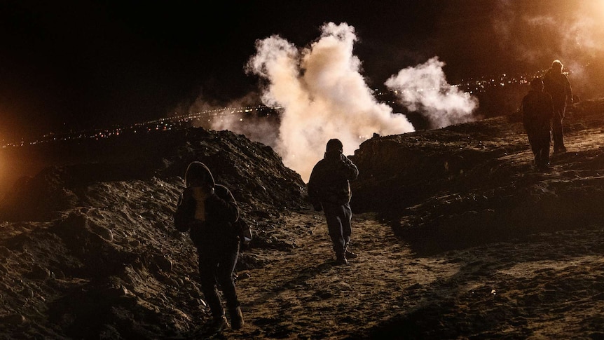 Four people run, covering their faces, as tear gas rises behind them at night time.