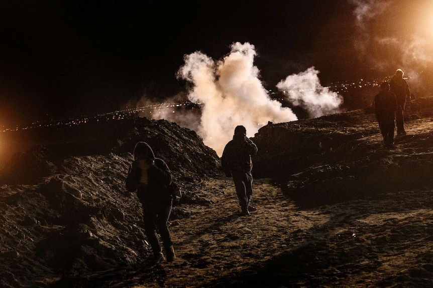 Four people run, covering their faces, as tear gas rises behind them at night time.