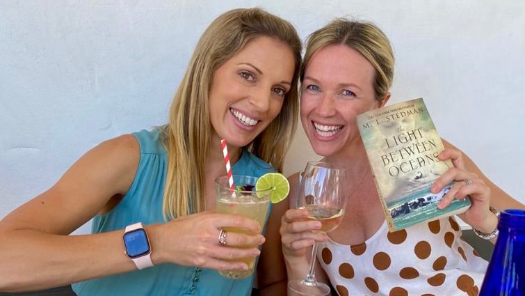 Two young women smiling, one holding a cocktail and the other holding a book