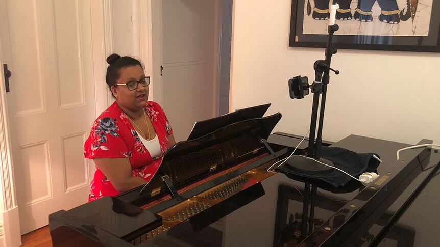 A woman sits at a piano with a phone live streaming her performance.
