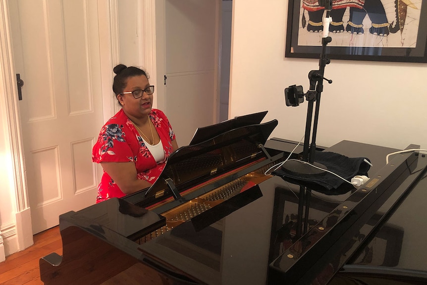 A woman sits at a piano with a phone live streaming her performance.