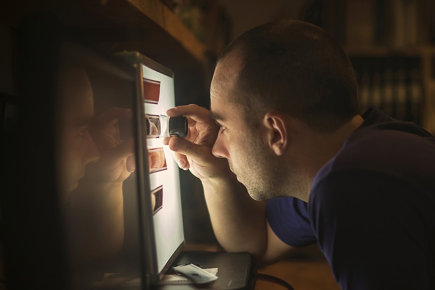 You view a man in a dark room whose face is lit up by a light box, as he uses a magnifying glass to look at 35mm film.