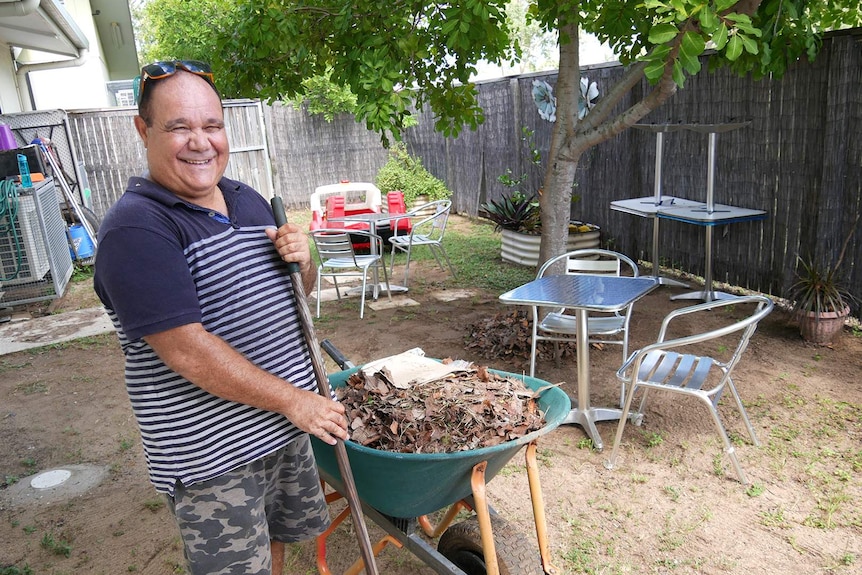 A man stands beside a wheelbarrow of leaves, he is using a rake.