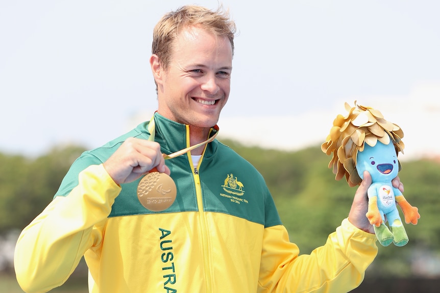 Curtis McGrath holds his medal and the Rio Paralympics mascot while on the podium and smiles.