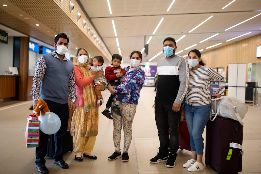 An extended family of two couples with a child each and a grandmother pose for a photo in an airport