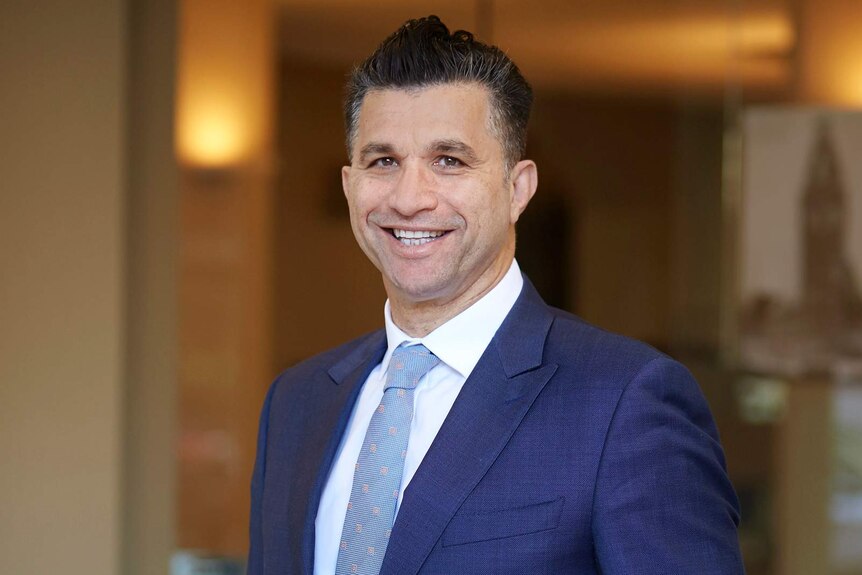 A mid shot of a middle-aged man in a blue suit as he poses for a photo with a big smile on his face.