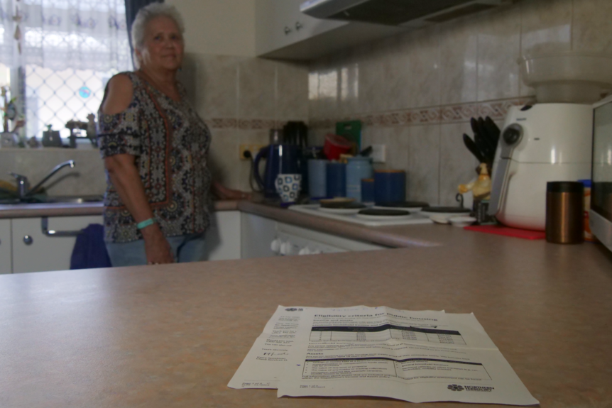 An elderly woman stands in her kitchen, with some paperwork visible in the foreground.