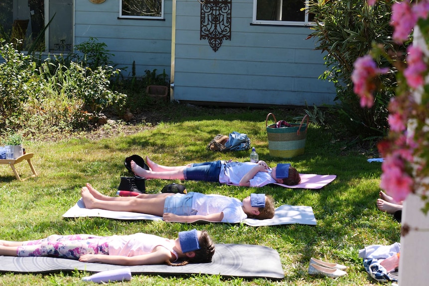 Young girls lying on yoga mats on a sunny lawn outside, with an old house in the background.
