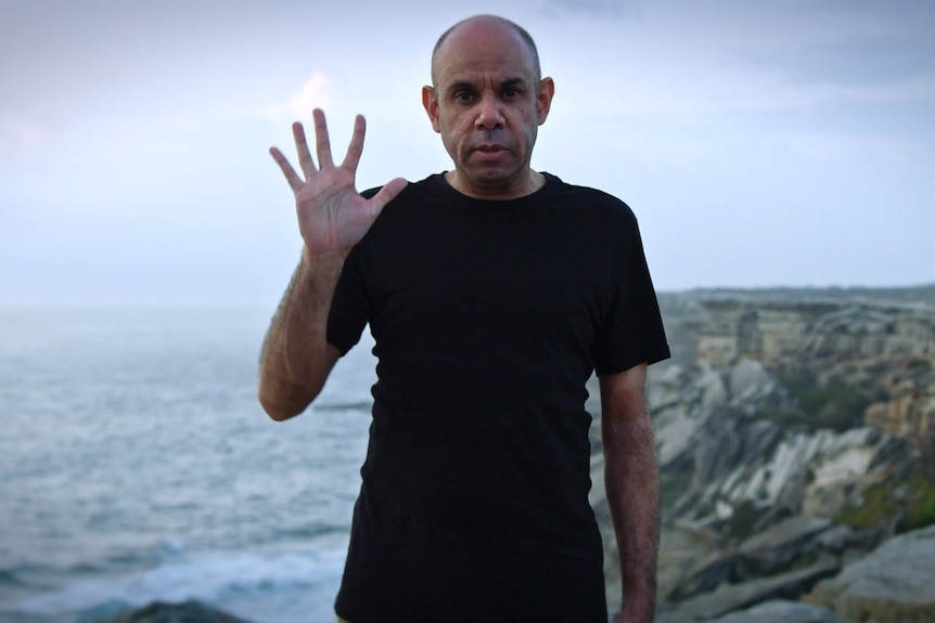 Steven Oliver standing on a cliff, ocean in the background, in a still from film Looky Looky Here Comes Cooky