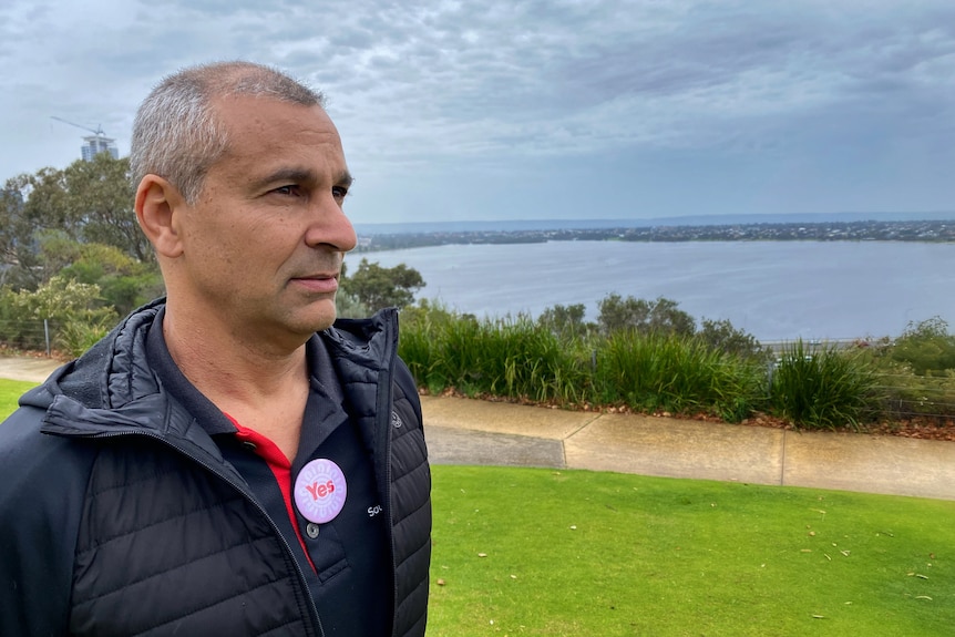 A man with short grey hair wearing a Yes badge and jacket looks out with water behind him.