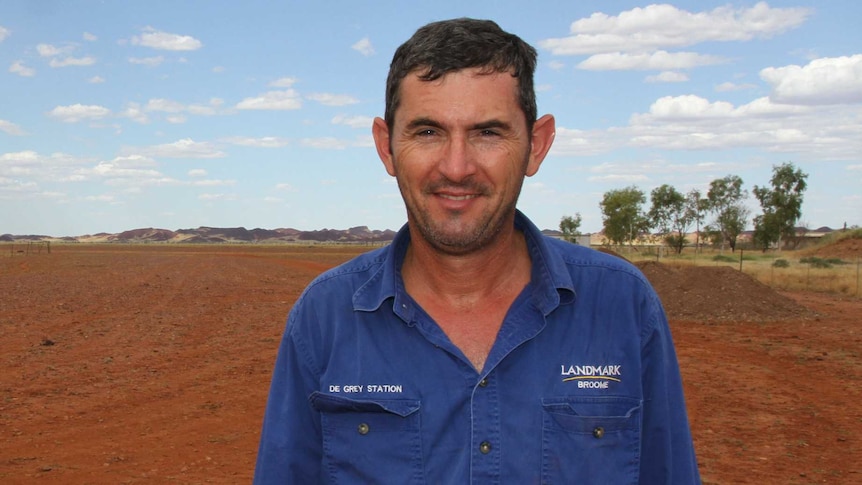 A man stands smiling in a red dirt paddock with hills behind him in the distance
