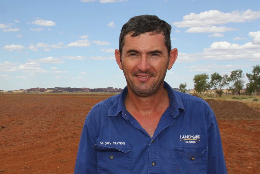 A man stands smiling in a red dirt paddock with hills behind him in the distance