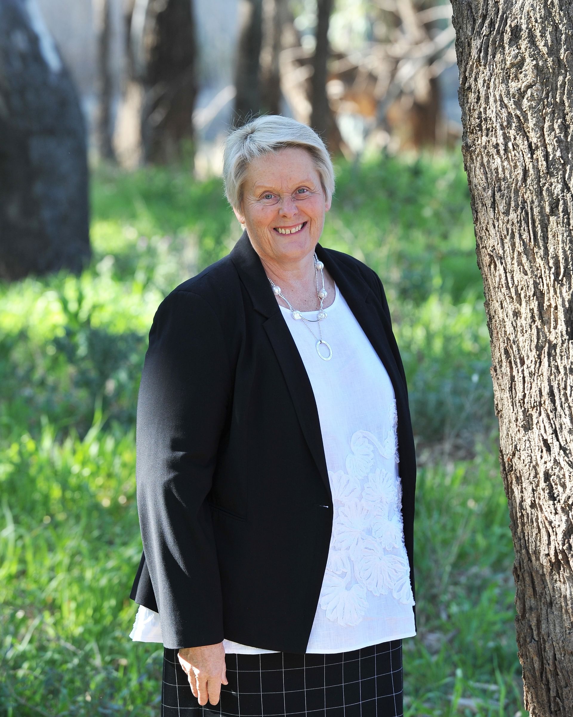 Woman with short hair, wearing a white shirt and black jacket, smiling standing among trees