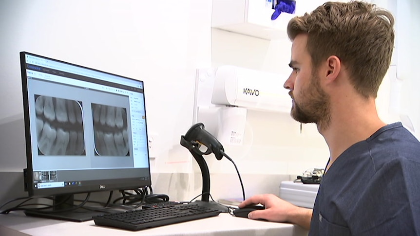 A man seated at a desk looking at X-rays of teeth on a computer screen.