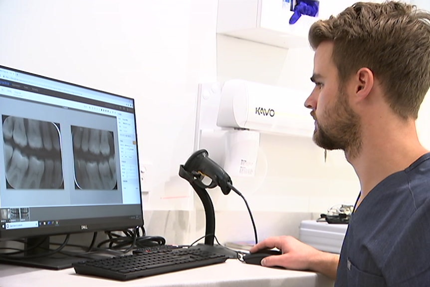 A man seated at a desk looking at X-rays of teeth on a computer screen.