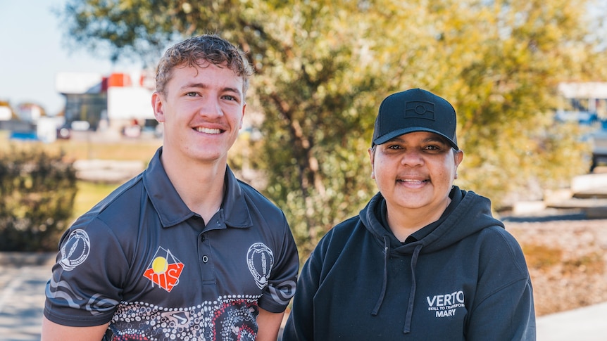 A young man in a shirt with Indigenous art on it standing next to a woman in a black jumper and hat. 