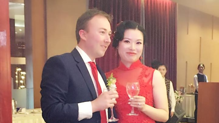 Adam Brown, in a suit, and Chen Cheng, in a red dress, hold drinks at a wedding reception