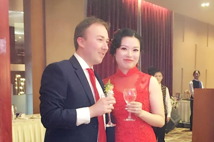 Adam Brown, in a suit, and Chen Cheng, in a red dress, hold drinks at a wedding reception