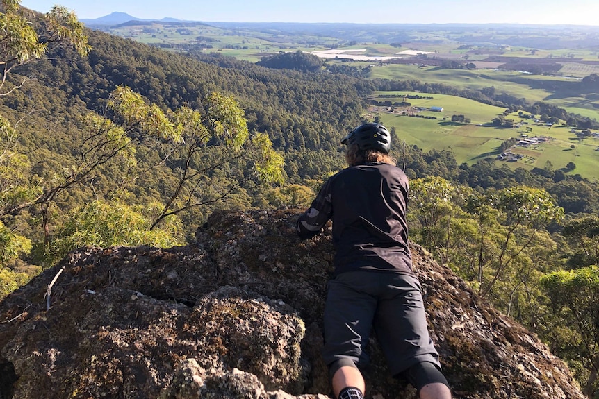A cyclist lays on a boulder to take in a view across farmland to a distant peak