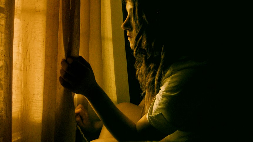 A woman looks through a window from a dimly lit room.