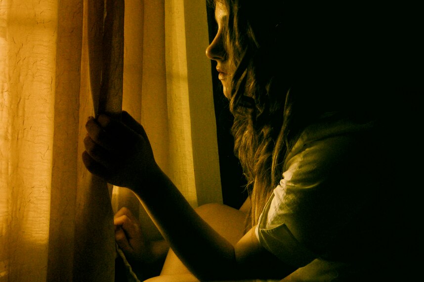A woman looks through a window from a dimly lit room.