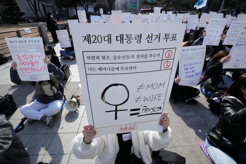 People sit cross-legged on the ground holding placards with Hangul script and feminist icons