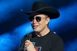 Elon Musk wearing a cowboy hat and sunnies, speaking with a microphone on stage