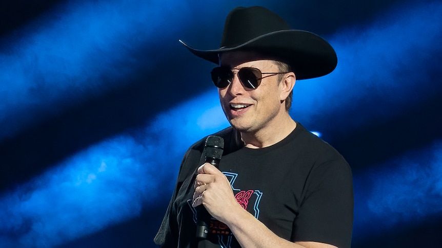 Elon Musk wearing a cowboy hat and sunnies, speaking with a microphone on stage