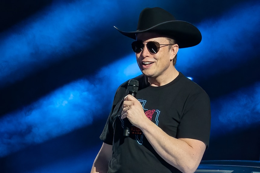 Elon Musk wearing a cowboy hat and sunnies, speaking with a microphone on stage