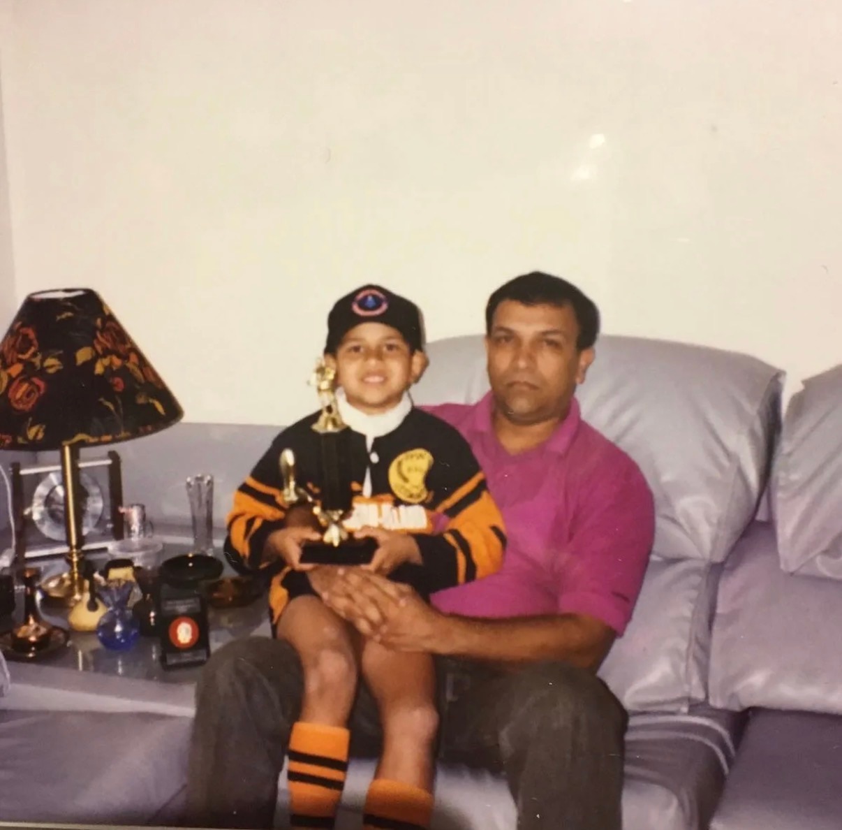 Usman Khawaja as boy sitting on his father's lap on couch. He holds several trophies. The boy is smiling while the father isn't.