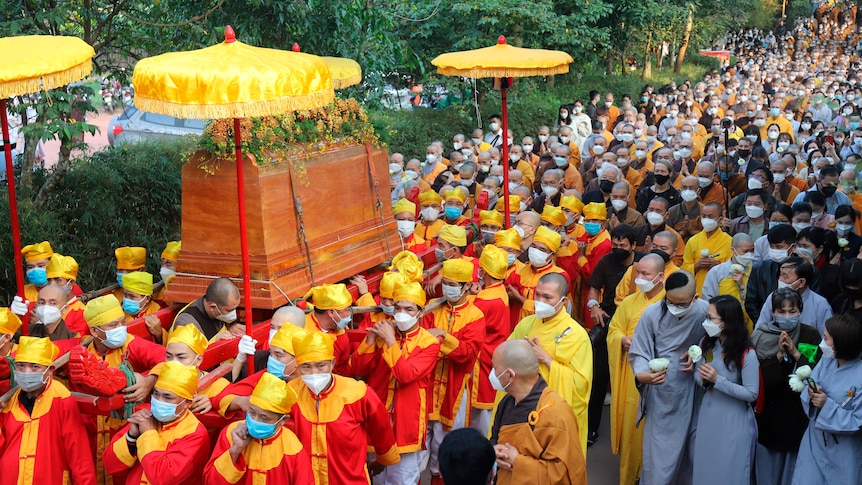 A huge procession of buddhist monks wearing bright red and yellow clothes carry a casket.