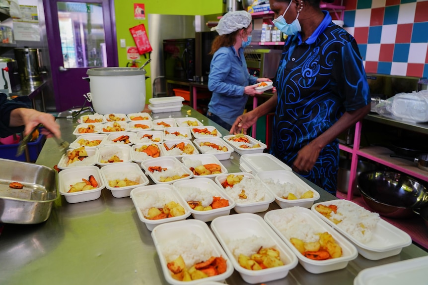 Two women in a commercial kitchen prepare dozens of meal trays filled with potatoes and rice.