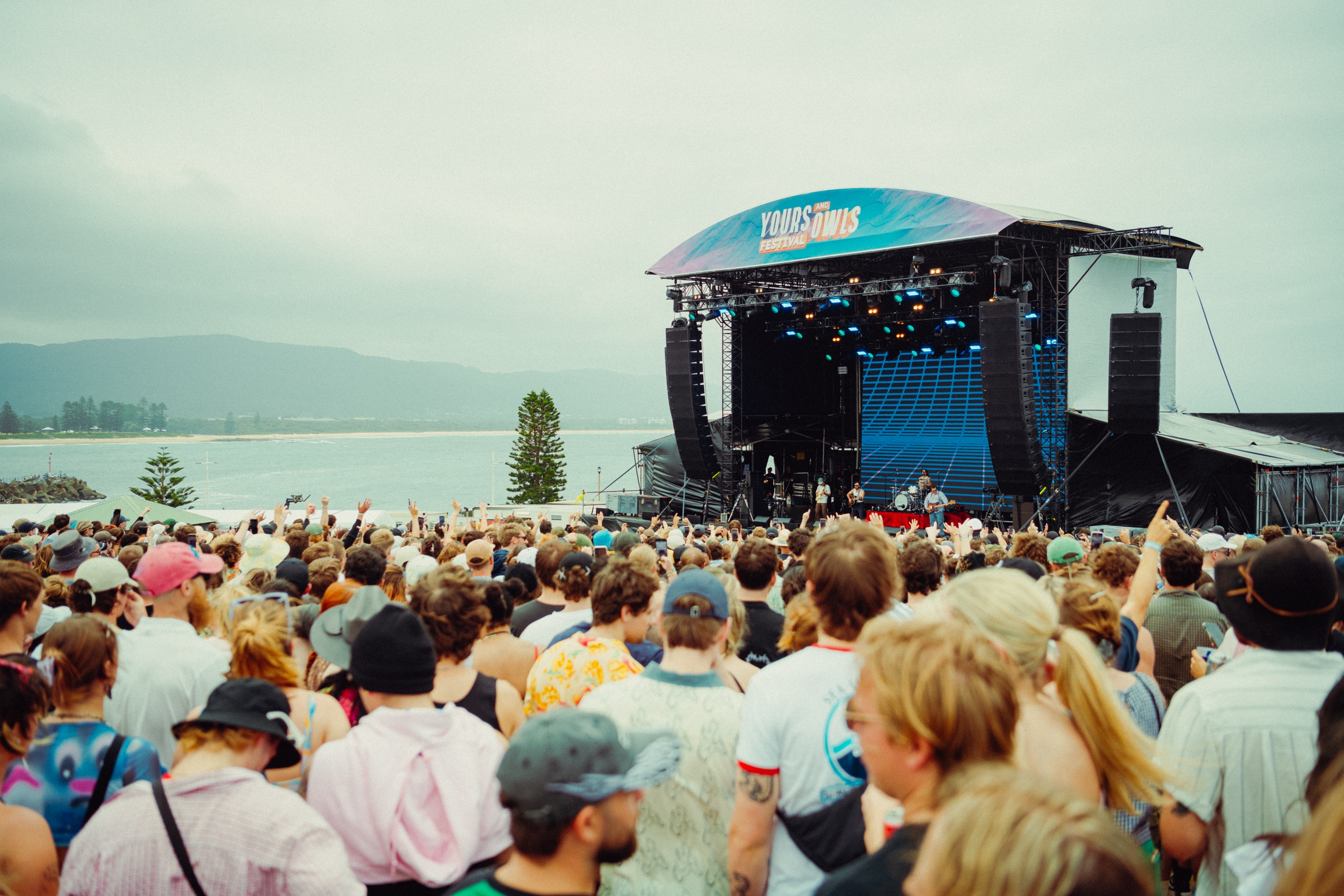 A crowd faces a stage on an overcast day with a beach line in the background