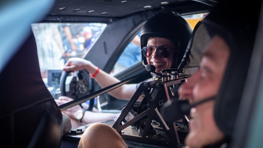 A woman and a man sit in a car with helmets and headsets. They are smiling.