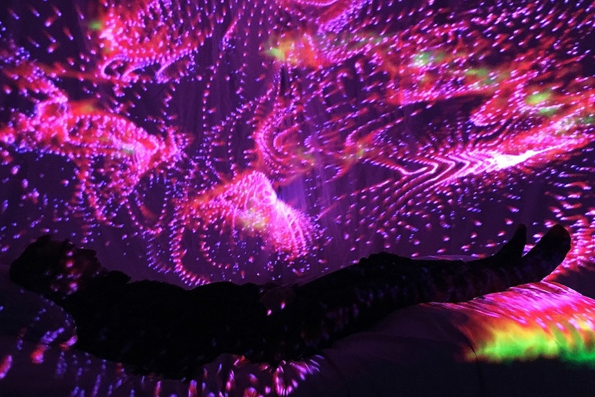 Participant lies in interactive bed wearing VR headset, surrounded by kaleidoscopic visuals.