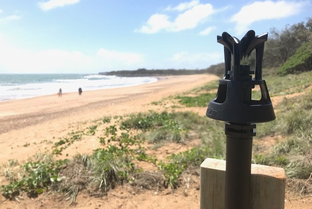 A black sprinkler head against a timber pole with a curved beach and vegetation-covered sand dune in the background.