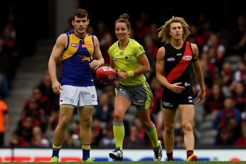 A woman in an umpire uniform carries a football between two male football players