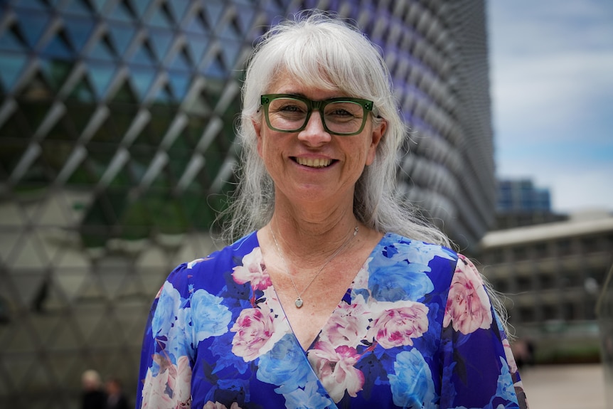 a woman with long grey hair with a fringe and green glasses smiles broadly