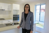 A professionally-dressed woman in a grey blazer stands on the property of a one-storey suburban home