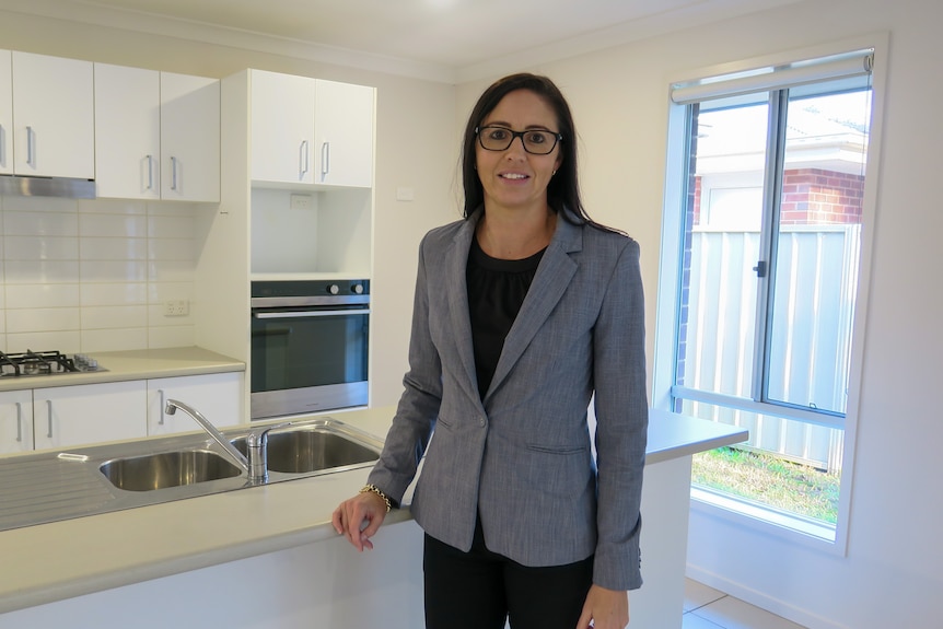 A professionally-dressed woman in a grey blazer stands on the property of a one-storey suburban home