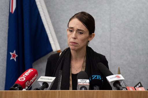 Jacinda Ardern's face is drawn and saddened as she stands at a podium with microphones pointed at her.