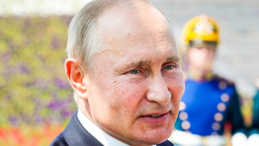 A close up of Vladimir Putin's face with a uniformed soldier in the background