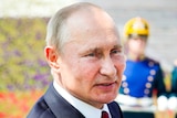 A close up of Vladimir Putin's face with a uniformed soldier in the background