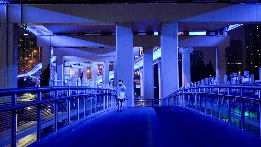 A person in a mask walks over a bridge bathed in blue light at night in Shanghai