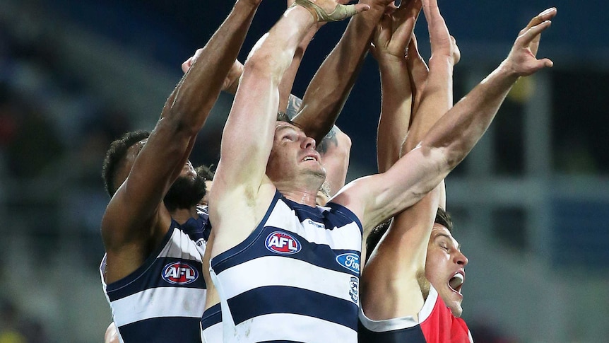 Male AFL players leap up in a pack with their arms stretched above the heads trying to mark the ball.