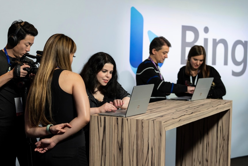 Four people standing at a desk, using two laptops between them. A man with a camera and a logo for Microsoft's Bing behind them.