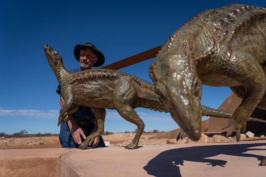 A man in a hat stands behind two small dinosaur statues on a sunny day in the outback.